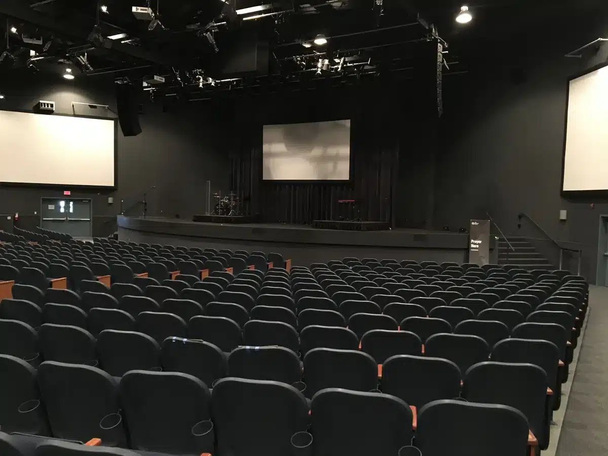 A packed IMAX theater auditorium with rows of empty seats under dramatic ceiling lighting, a single spotlight illuminating the massive curved screen, conveying the scale and grandeur of the premium format venue
