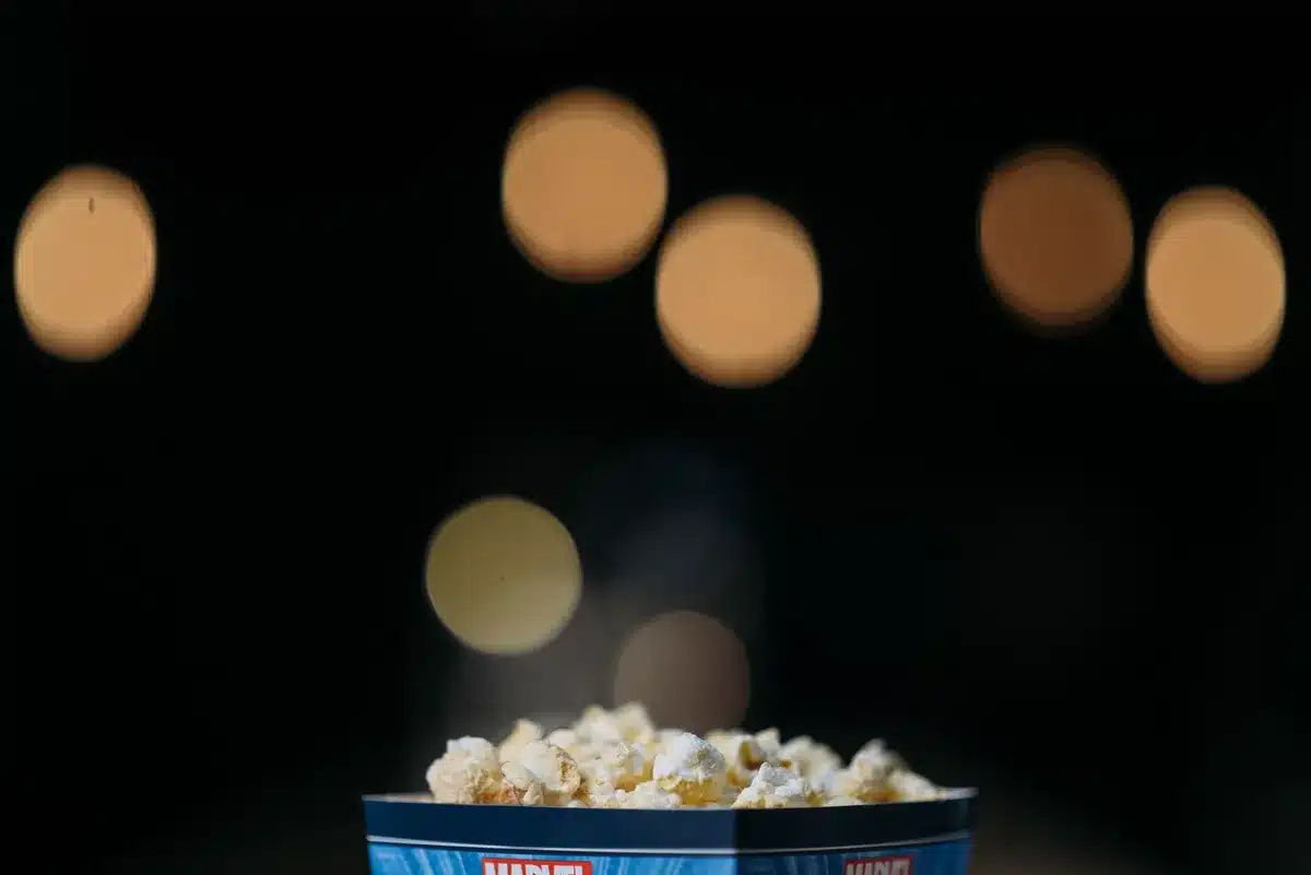 A packed movie theater lobby with crowds of moviegoers in casual attire, some holding tickets and concessions, under bright marquee lighting with the film title prominently displayed.