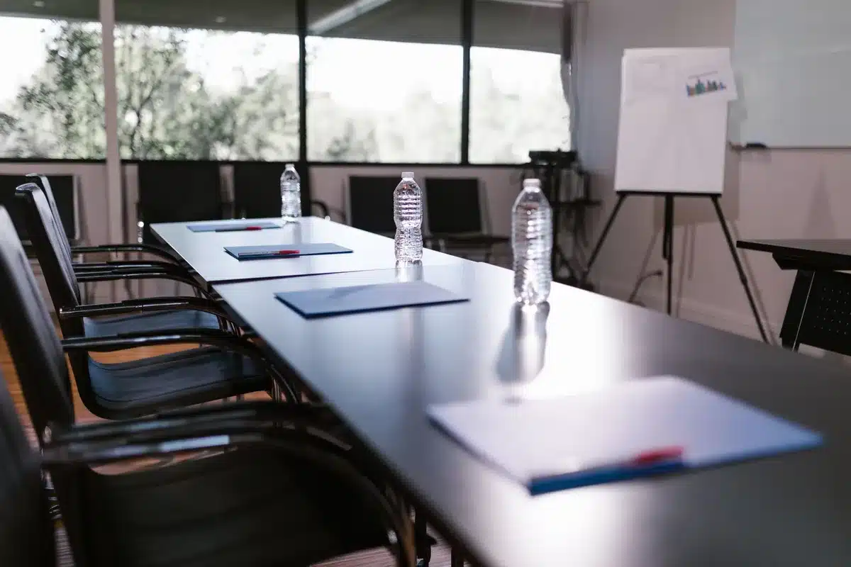 An empty modern boardroom with a single executive chair at the head of a glass table, sunlight streaming through floor-to-ceiling windows overlooking a corporate campus, symbolizing transition and legacy.