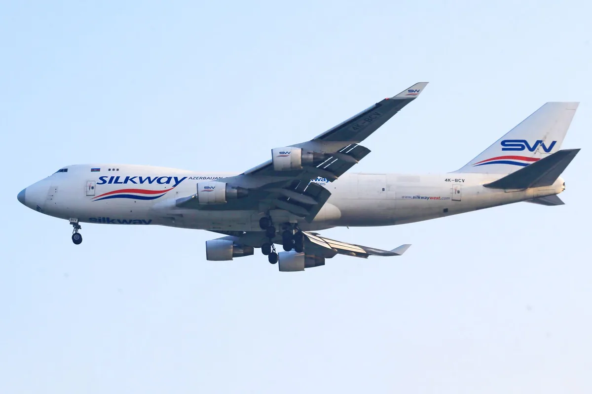 A massive four-engine jumbo jet in white livery descending toward a runway under clear skies, its landing gear extended, with terminal buildings visible in the distance below.