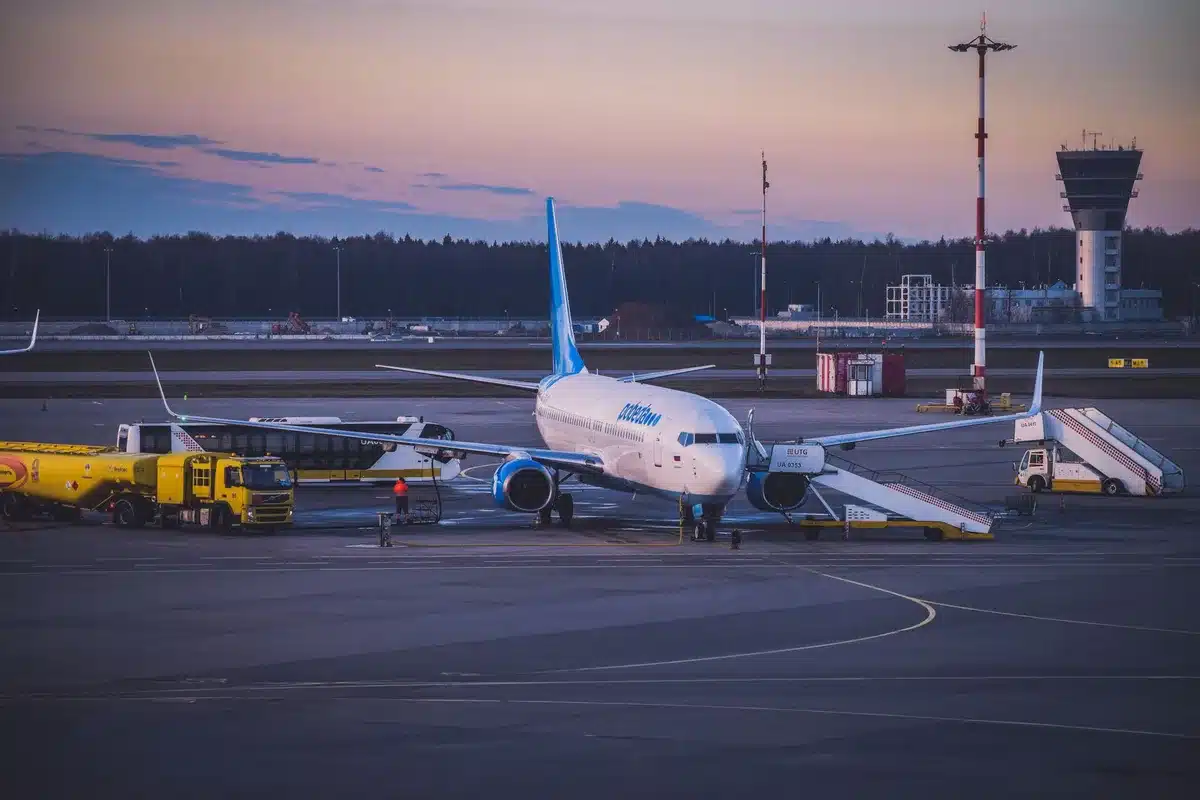 A wide-angle view of a commercial aircraft on a tarmac with ground service vehicles positioned nearby, the airport terminal visible in the background under bright daylight.