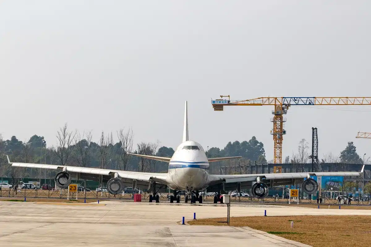 A massive unbranded white jumbo jet on a tarmac under bright floodlights, nose pointed toward the camera, fuselage gleaming with precision and scale, technicians in safety gear visible near the landing gear
