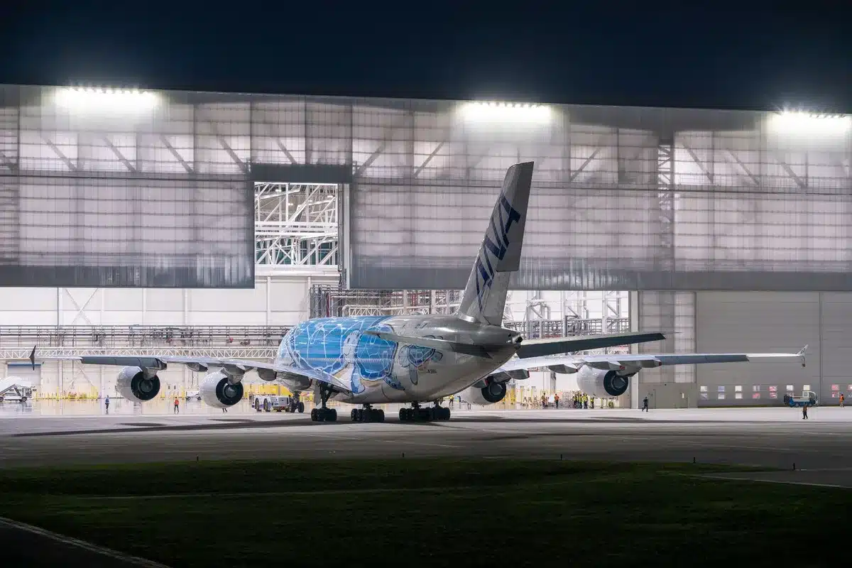 A sleek 747-8 aircraft fuselage under bright hangar lights, partially assembled with visible wiring and structural components, technicians in hard hats working nearby, industrial scaffolding surrounding the frame