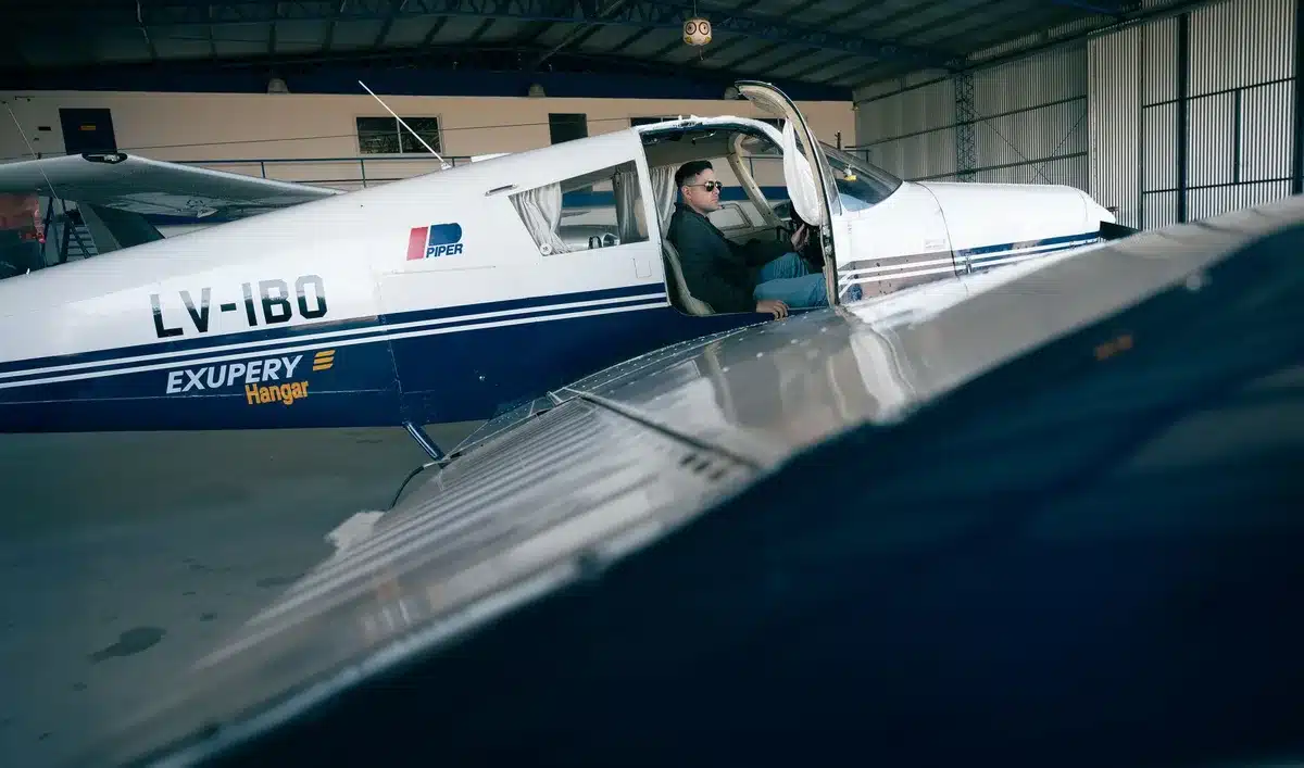 An empty, dimly lit aircraft hangar interior with overhead lights casting long shadows, a single work station visible in the distance, no personnel present, conveying the scale and scope of aerospace manufacturing