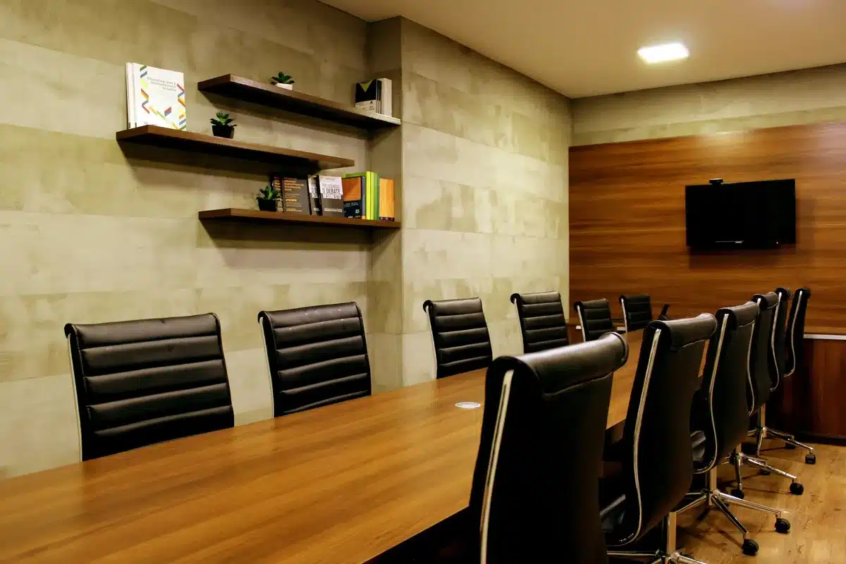 Empty boardroom with a single vacant executive chair at the head of a polished conference table, soft natural light from tall windows casting long shadows across dark wood, conveying loss and uncertainty