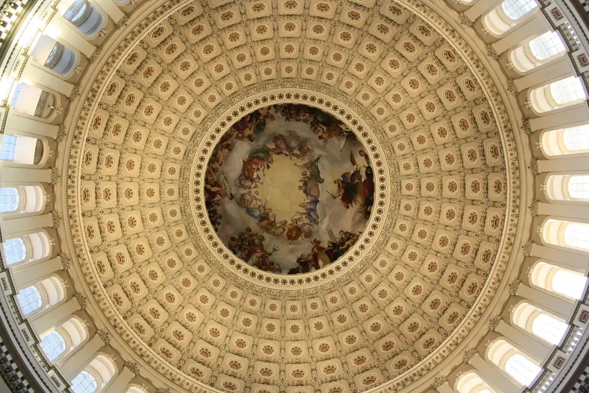 Capitol Rotunda interior with marble columns and domed ceiling, casket draped in flag on catafalque, candles and wreaths surrounding, solemn light filtering through skylights