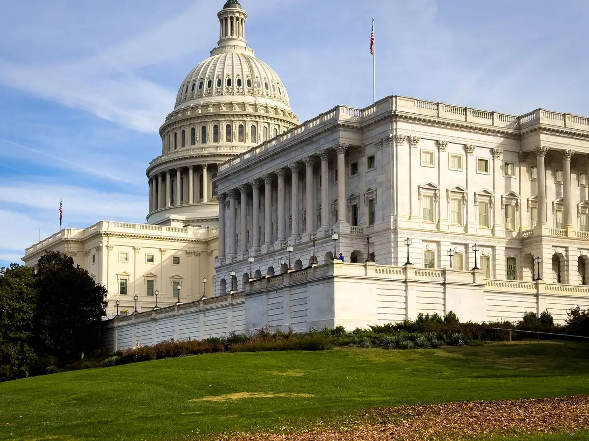 Capitol exterior at dusk with American flags at half-staff, building illuminated against evening sky, crowds of people gathered outside