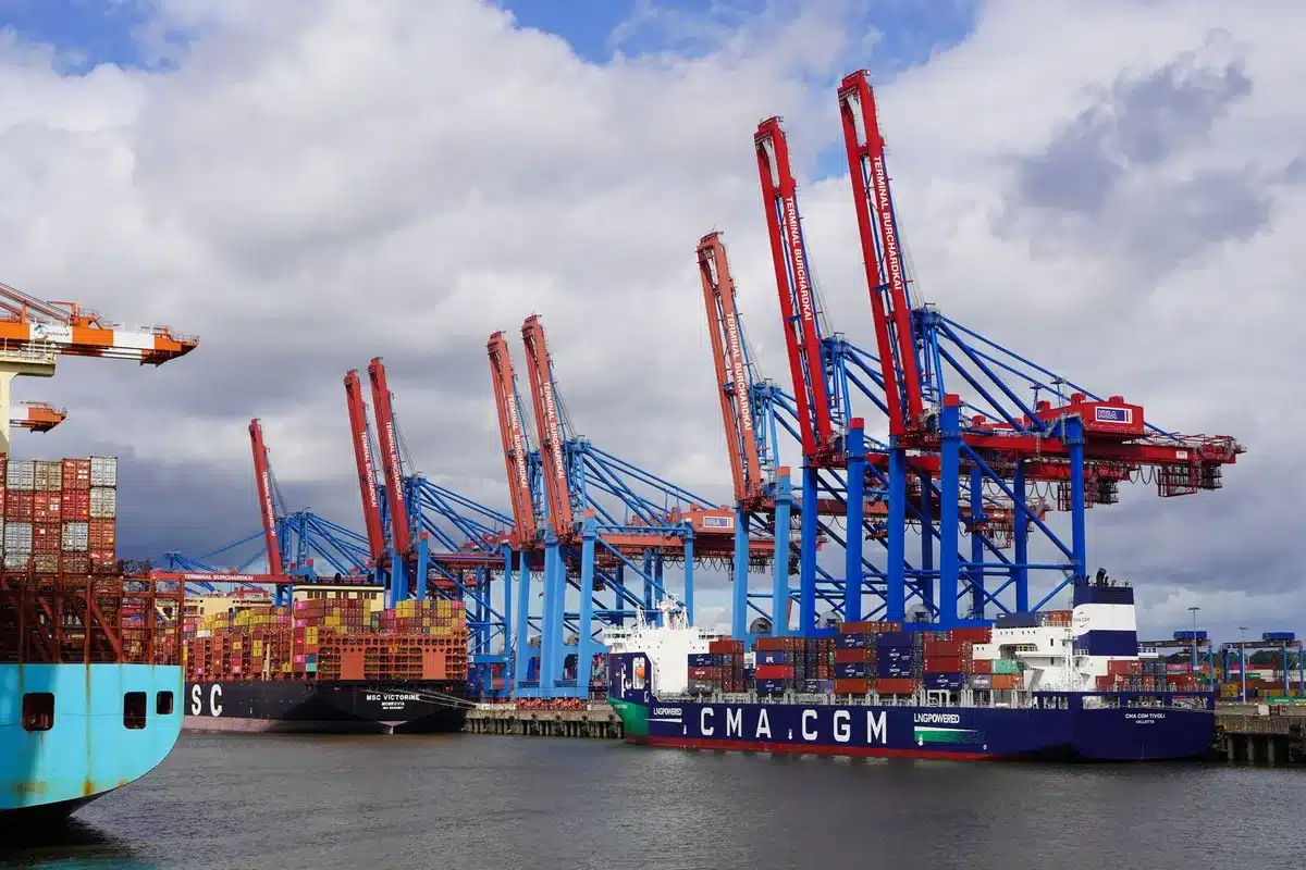 A shipping container at a port with tariff documents or trade paperwork visible nearby, industrial cranes in soft focus, suggesting cross-border commerce under strain.