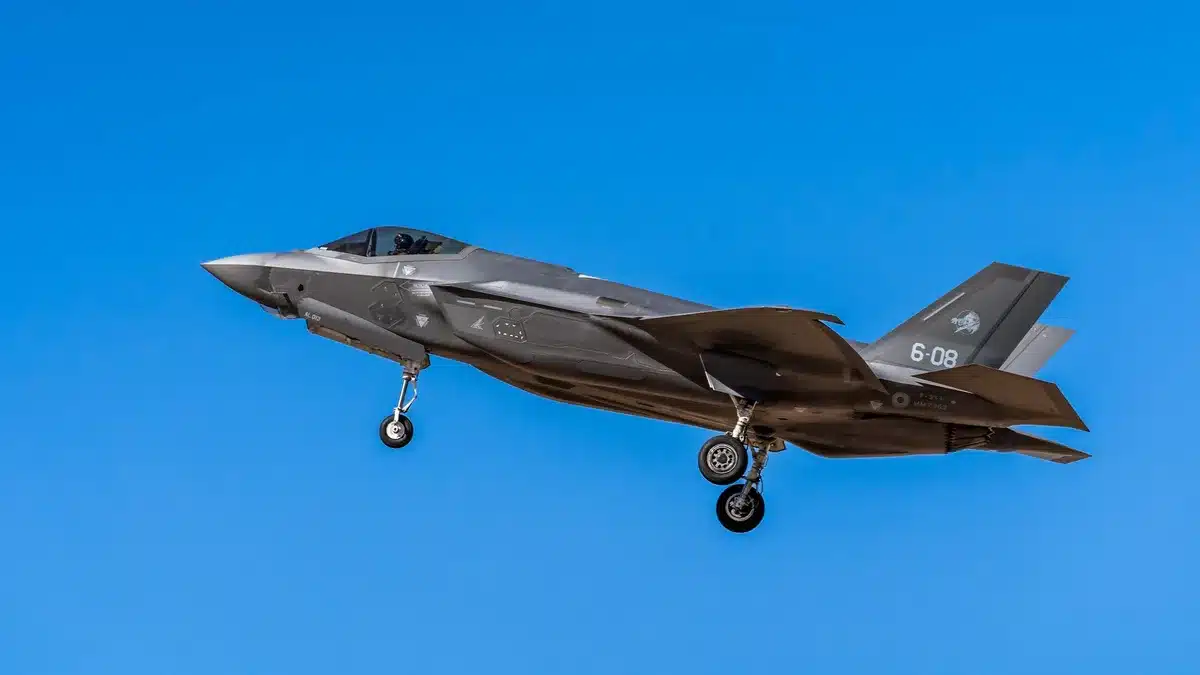 Military hangar with F-35B fighter jet on display, monarch in formal suit walking alongside uniformed service personnel, flags and aircraft in background, afternoon light
