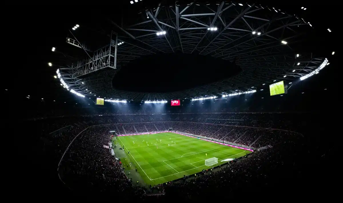 A stadium scoreboard displaying a beverage brand during a major sporting event, with cheering fans blurred in the foreground, colorful lighting and digital displays illuminating the venue atmosphere