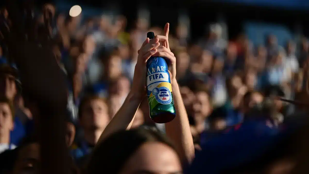 A beverage bottle with FIFA World Cup branding or stickers, held by hands in a celebratory gesture, with a crowd in a stadium background slightly out of focus