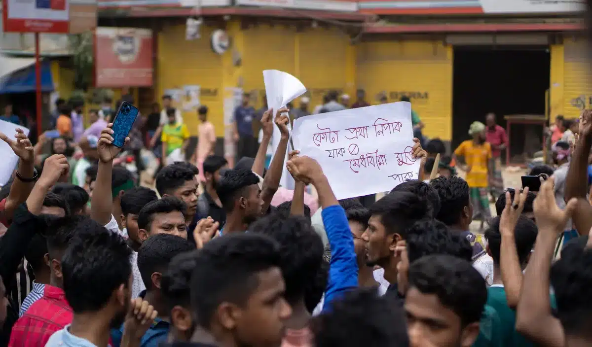 Crowded protest scene with thousands of anonymous students holding placards demanding accountability, faces obscured in motion, under bright sunlight at a public square, collective determination and youth energy visible