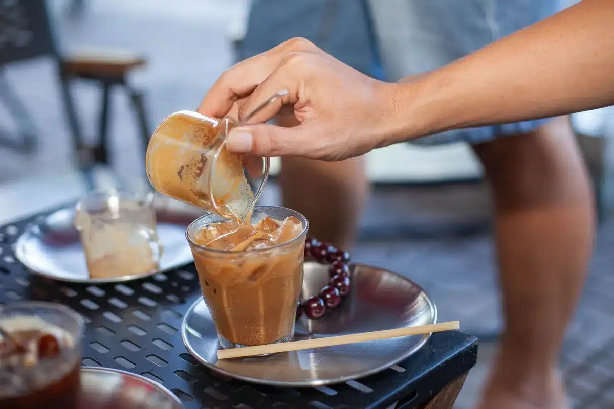 A 48-ounce beverage bucket filled with iced coffee, sitting upright with condensation on the exterior, photographed against a bright summer backdrop with beach or poolside setting visible in soft focus