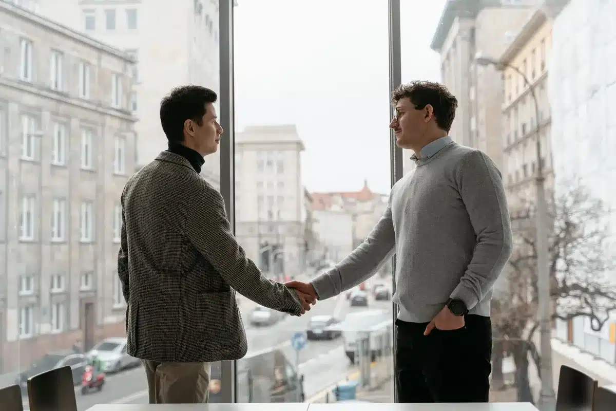 Two professionals in a modern office shaking hands across a desk, natural light streaming through windows, representing partnership and deal completion.