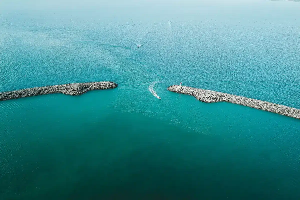 Narrow shipping lane bordered by rocky coastlines, oil tankers anchored in calm waters, international maritime traffic in suspension, tension visible in empty channels