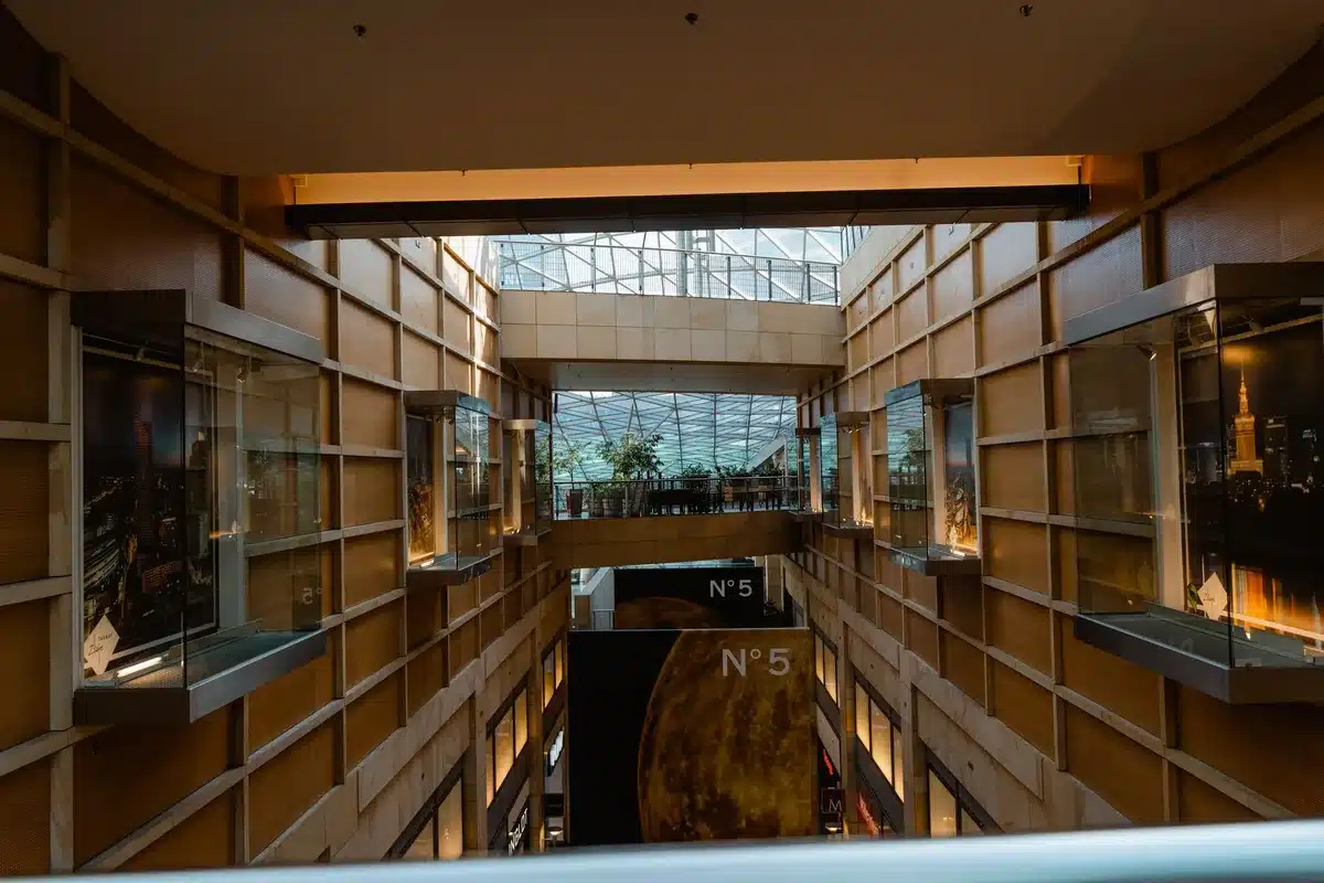A modern shopping mall interior with warm lighting and contemporary architecture, showing polished floors and sleek storefronts, with shoppers walking through a wide corridor lined with retail displays and ambient signage.