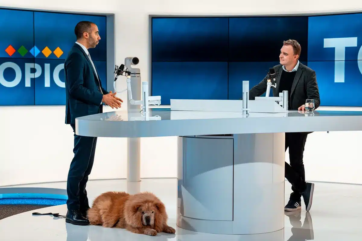 A television studio set with news desk and bright studio lighting, empty anchor chair facing camera, professional broadcast environment, cool blue and white tones