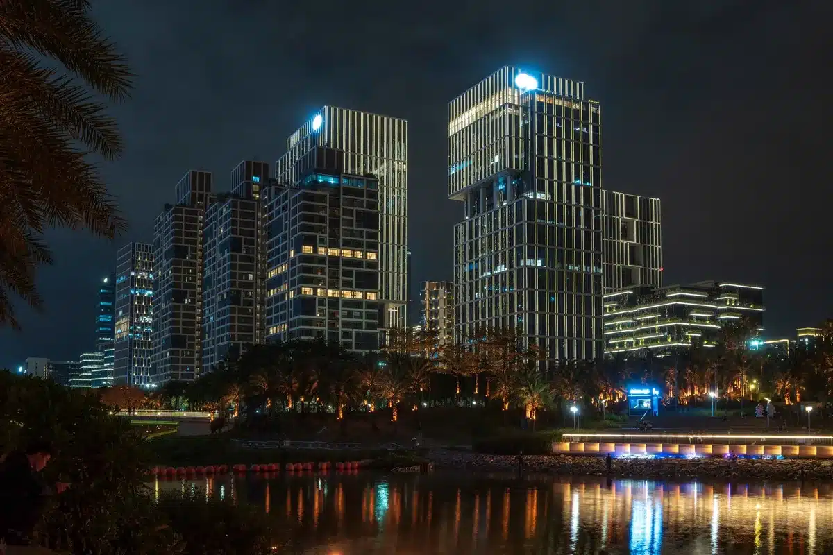 A high-rise office building with glass reflecting financial district skyline, evening light casting long shadows, a single lit window among darkened ones, empty streets below suggesting market closure