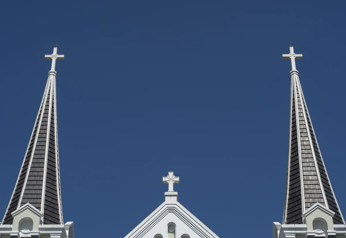 A simple wooden cross mounted on a white wall, with soft golden light emphasizing its form and shadow