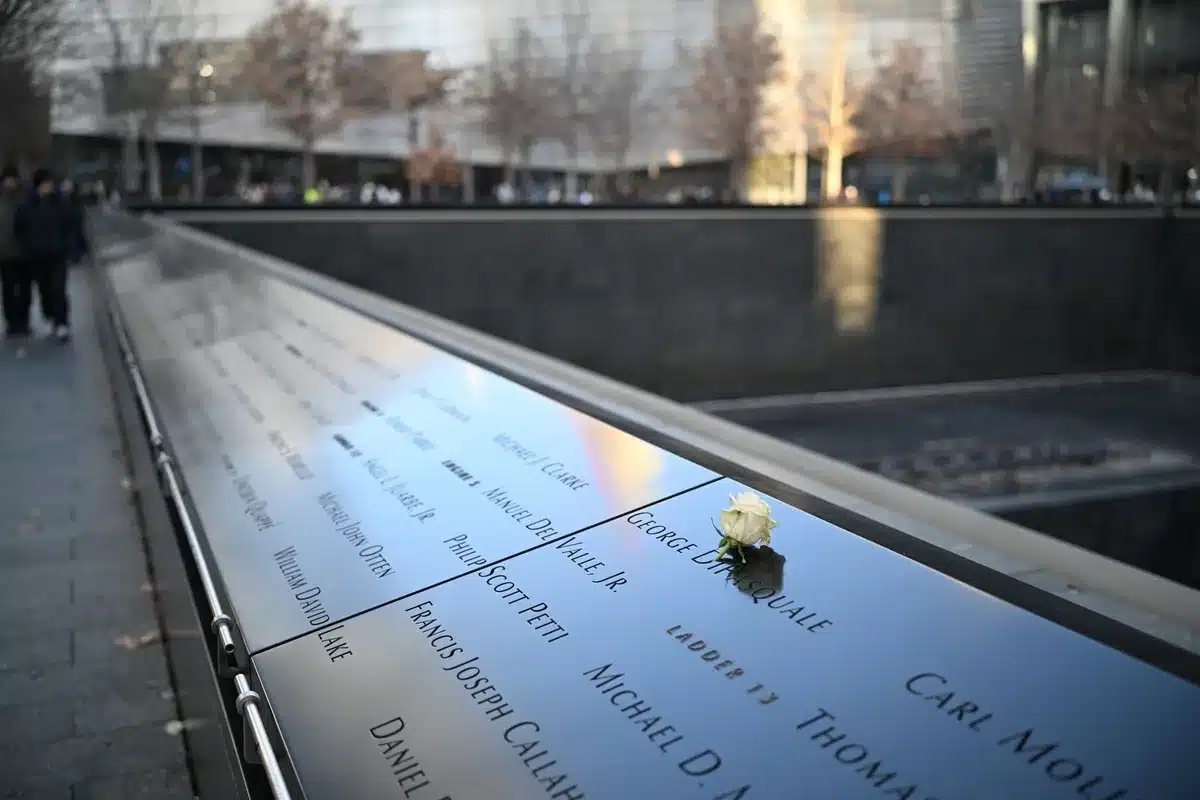 An empty memorial plaza at dawn, with the names of victims inscribed on stone, autumn light casting long shadows across the ground, a single wreath leaning against the wall, silence and stillness