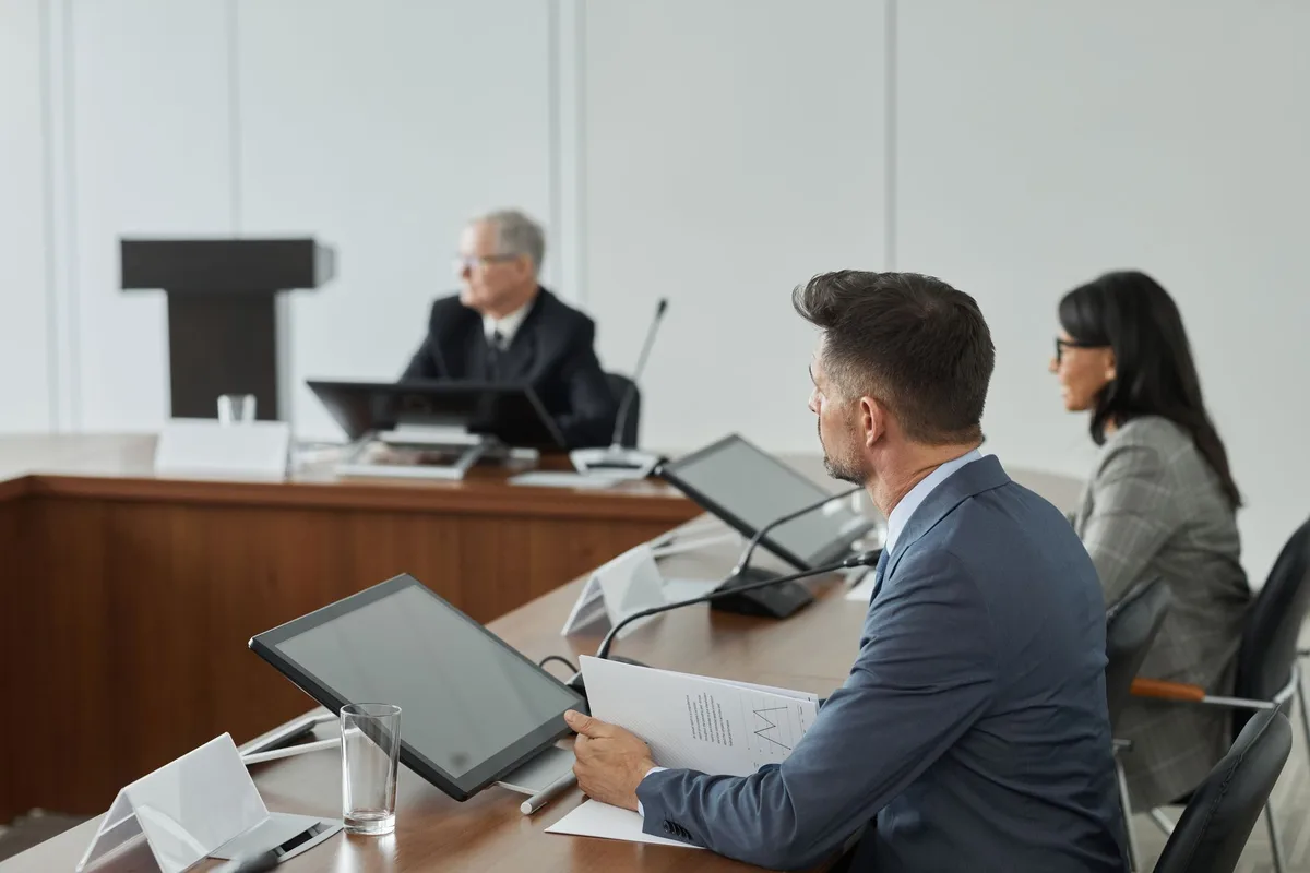 Political candidates and strategists gathered in a conference room, seated around a large table with notebooks and water glasses, having an animated discussion about campaign strategy and district outreach.