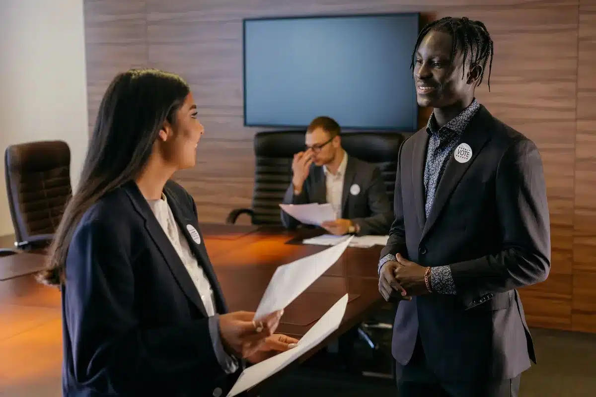 A diverse group of candidates and organizers gathered around a table in a modern conference room, reviewing campaign materials and discussing strategy, natural daylight streaming through large windows.
