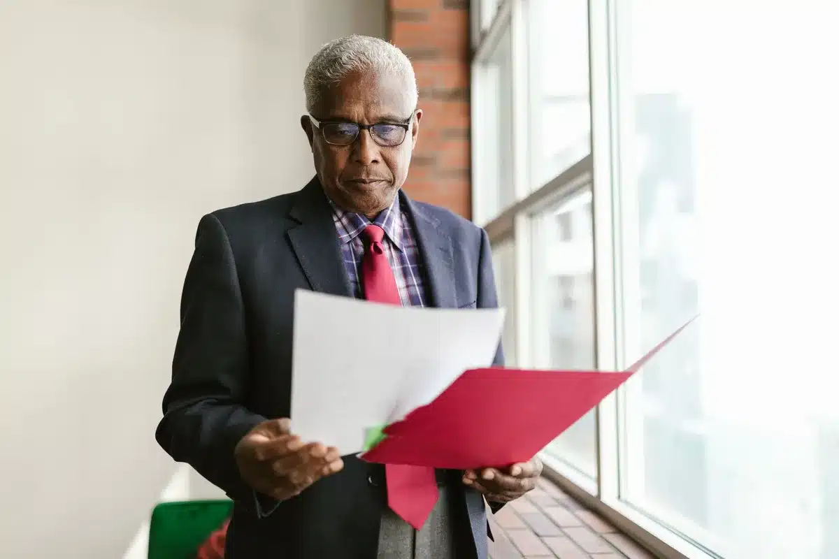A senior citizen reviewing medical paperwork at a kitchen table, glasses on, a cup of coffee nearby, morning light from a window illuminating documents about healthcare coverage.