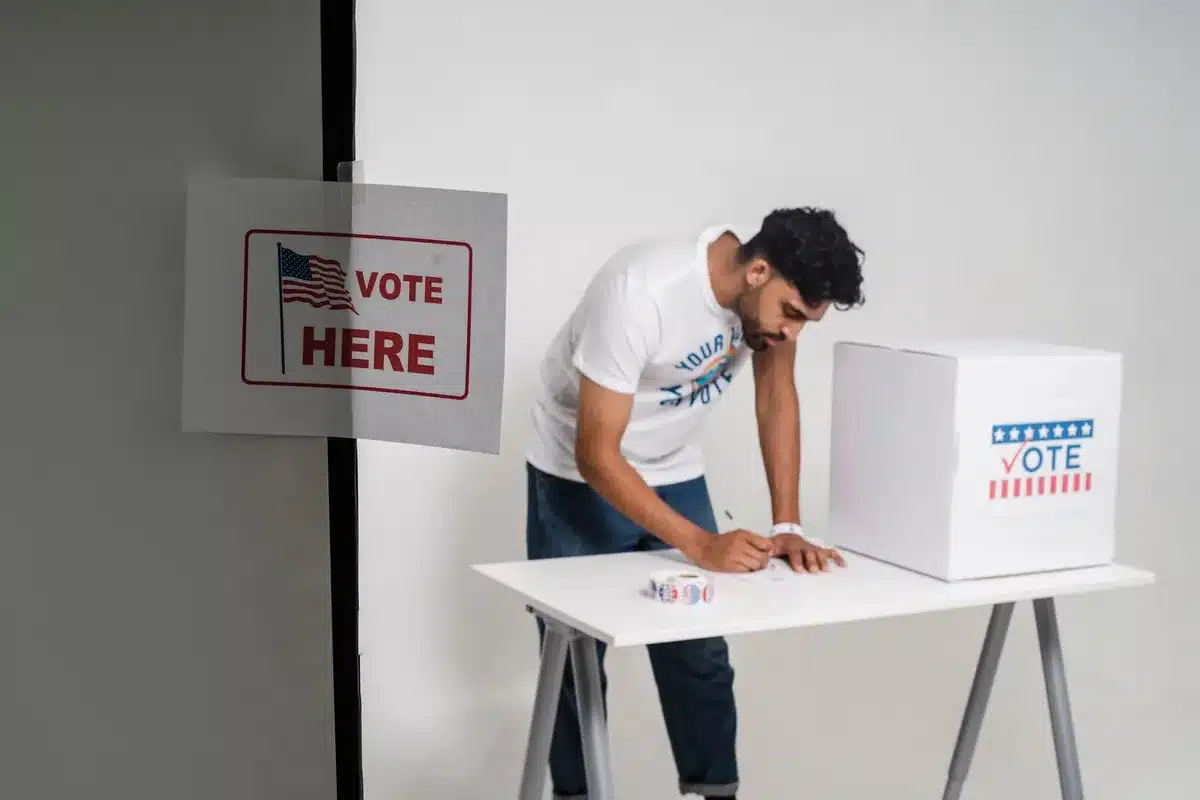 A ballot box with November 3, 2026 election date visible, with voting booths in soft focus behind it, representing the general election choice ahead