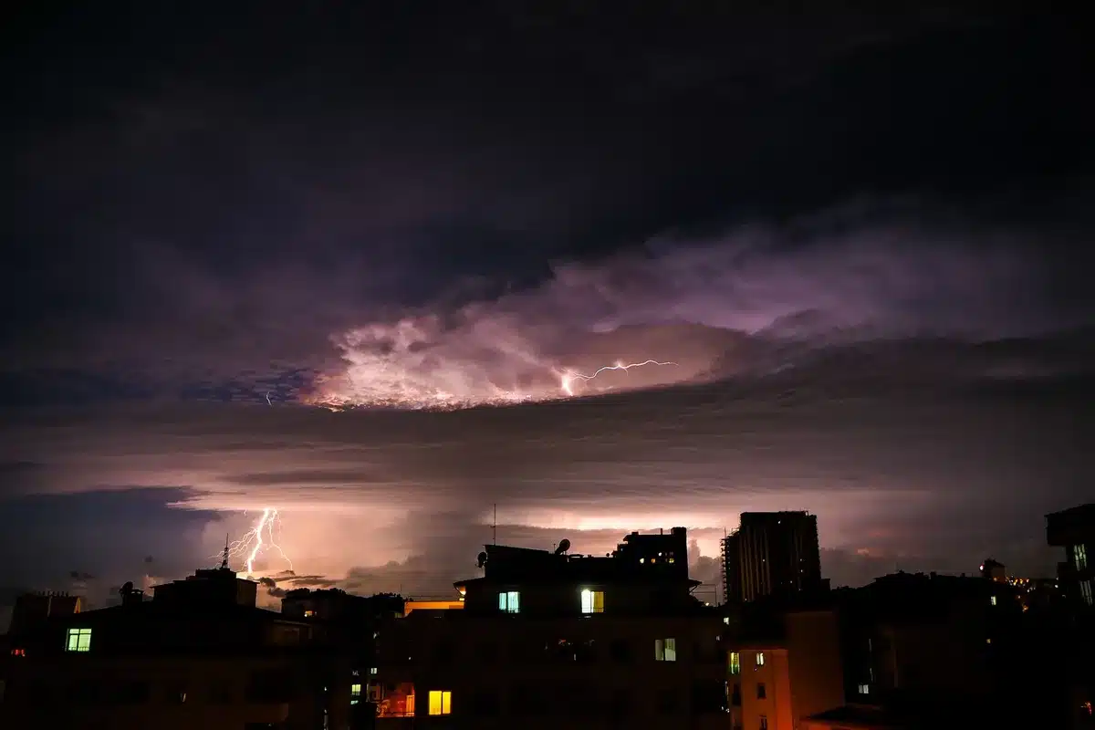 Dark storm clouds rolling over a residential neighborhood at dusk, lightning illuminating the sky in the distance, streetlights beginning to glow