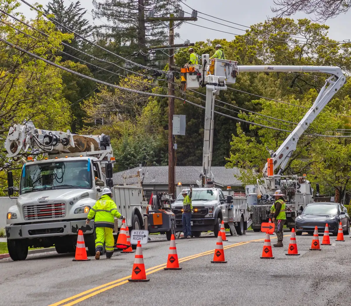 Crews in safety gear working on electrical lines and poles against a cloudy sky, utility trucks and equipment visible nearby.