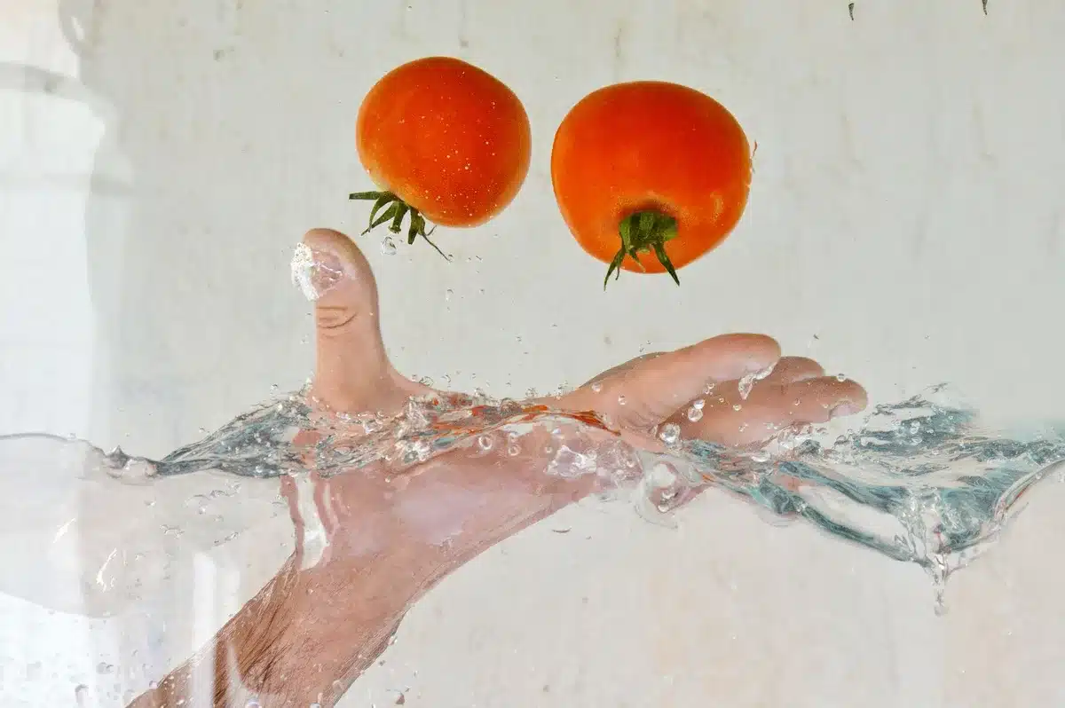 Fresh produce being washed under running water in a kitchen sink, water droplets visible, vegetables and herbs in a colander