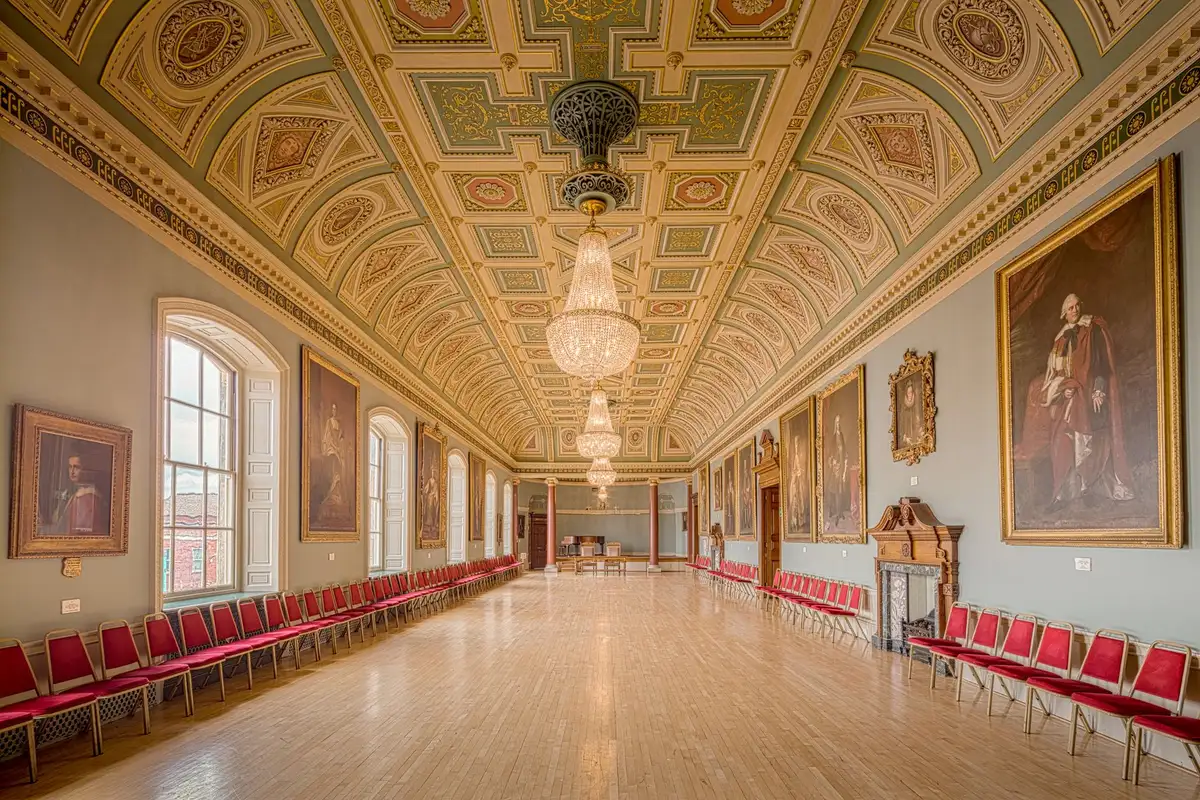 An empty formal state reception room with long polished wooden table, ornate chairs positioned on either side, national flags standing at attention in the background, soft golden light filtering through tall windows, a sense of diplomatic ceremony and negotiation suspended in time