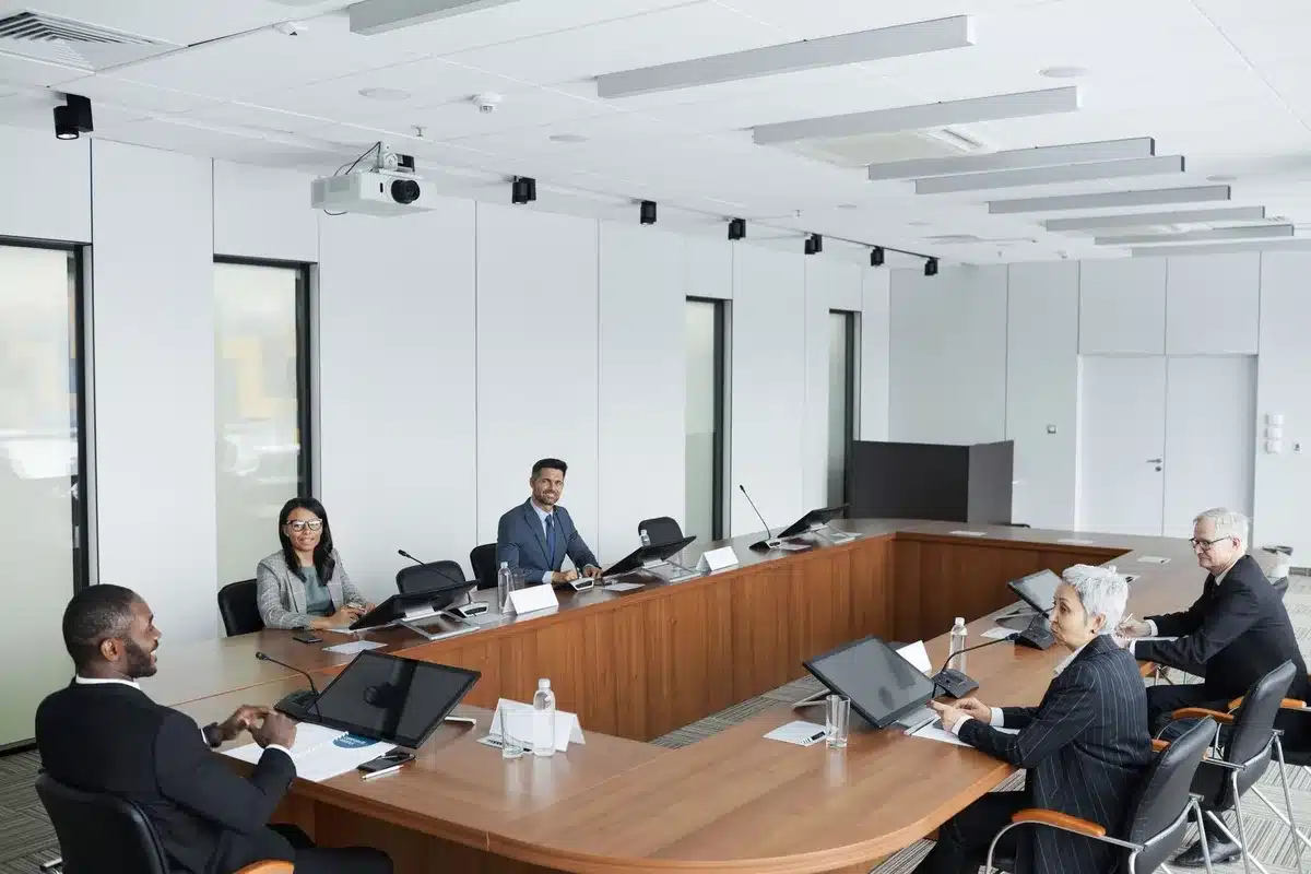 A diverse group of anonymous legal professionals in business attire seated around a conference table, some with hands raised in discussion, natural window light casting shadows across papers and legal documents, conveying collaborative decision-making without identifying individuals.