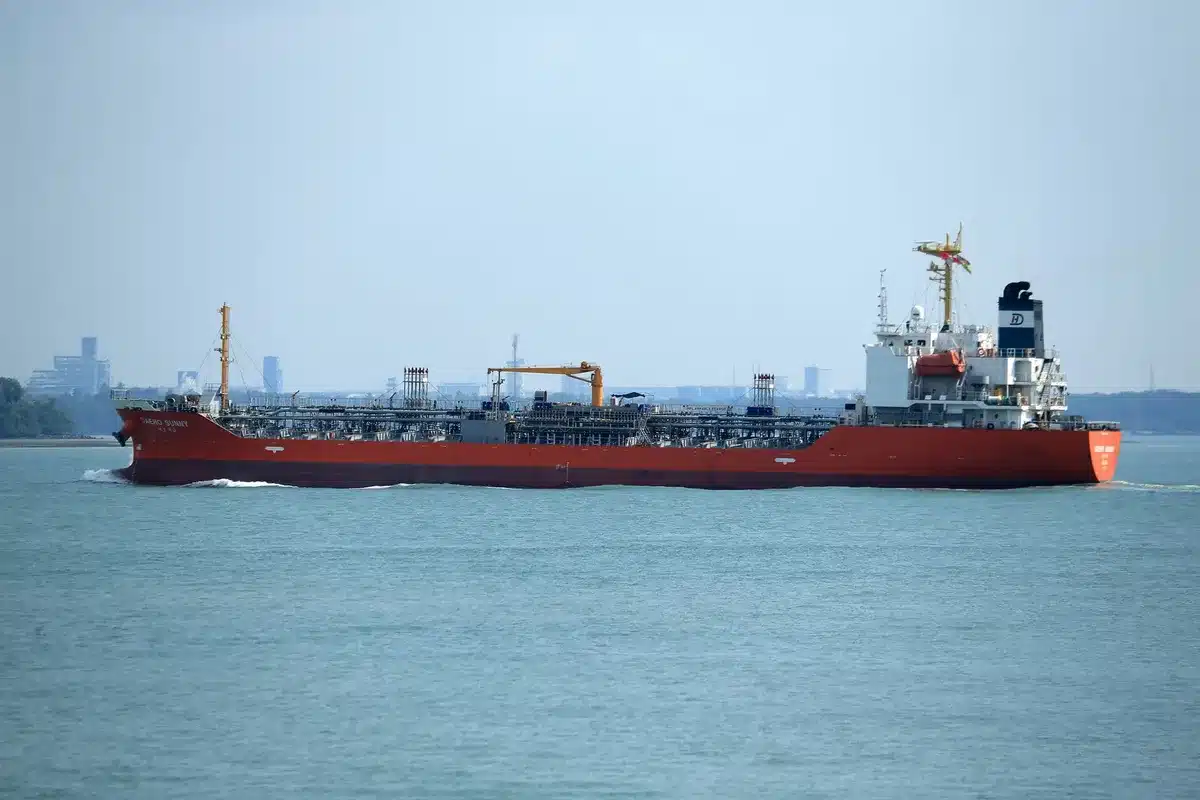 A crowded shipping lane with multiple cargo vessels lined up in narrow waters, some with running lights visible at dusk, oil tankers waiting to transit a contested strait
