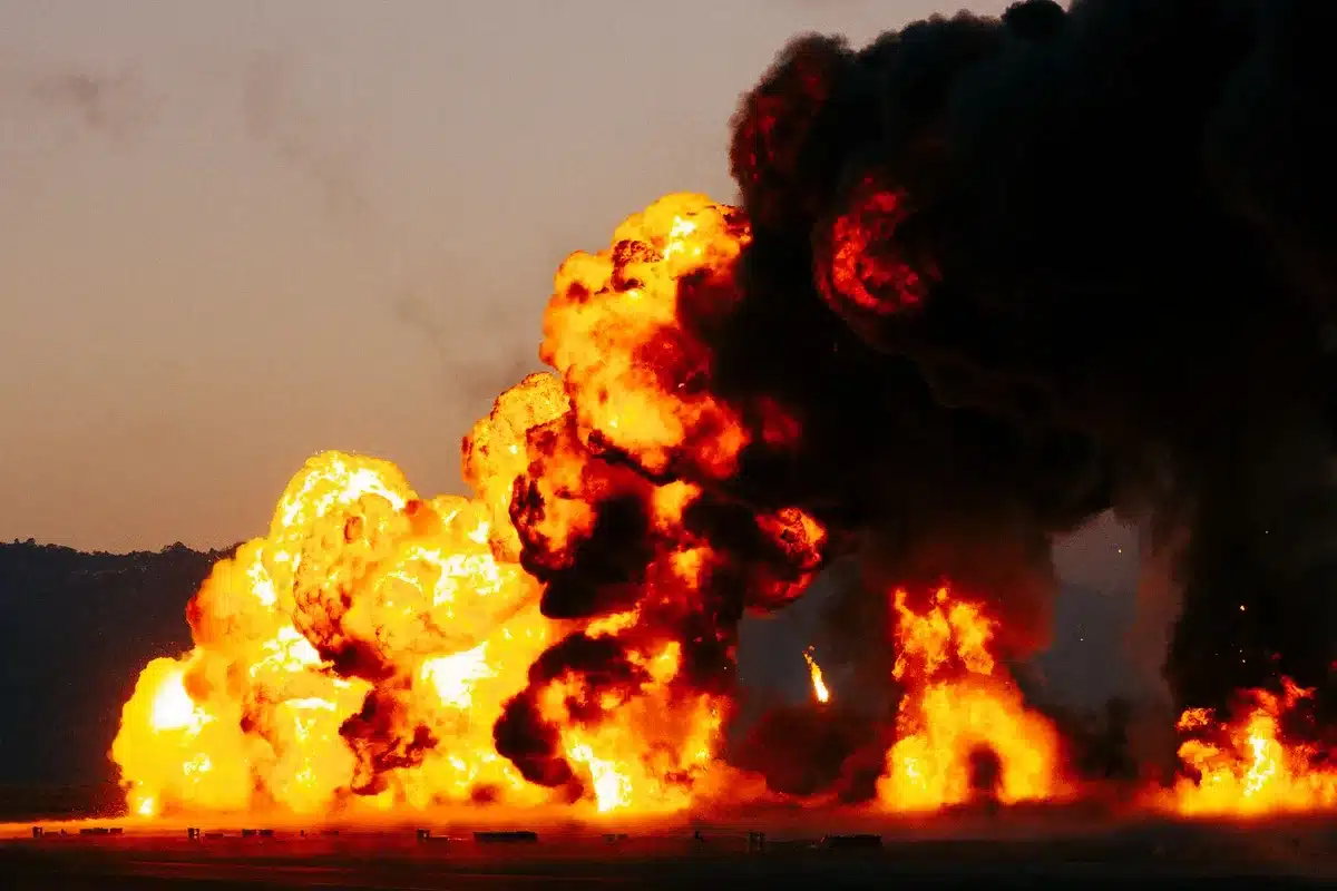 An oil tanker engulfed in flames on dark water, smoke billowing across the surface, a naval mine aftermath visible in the distance, the vessel listing to one side, emergency response vessels approaching through haze