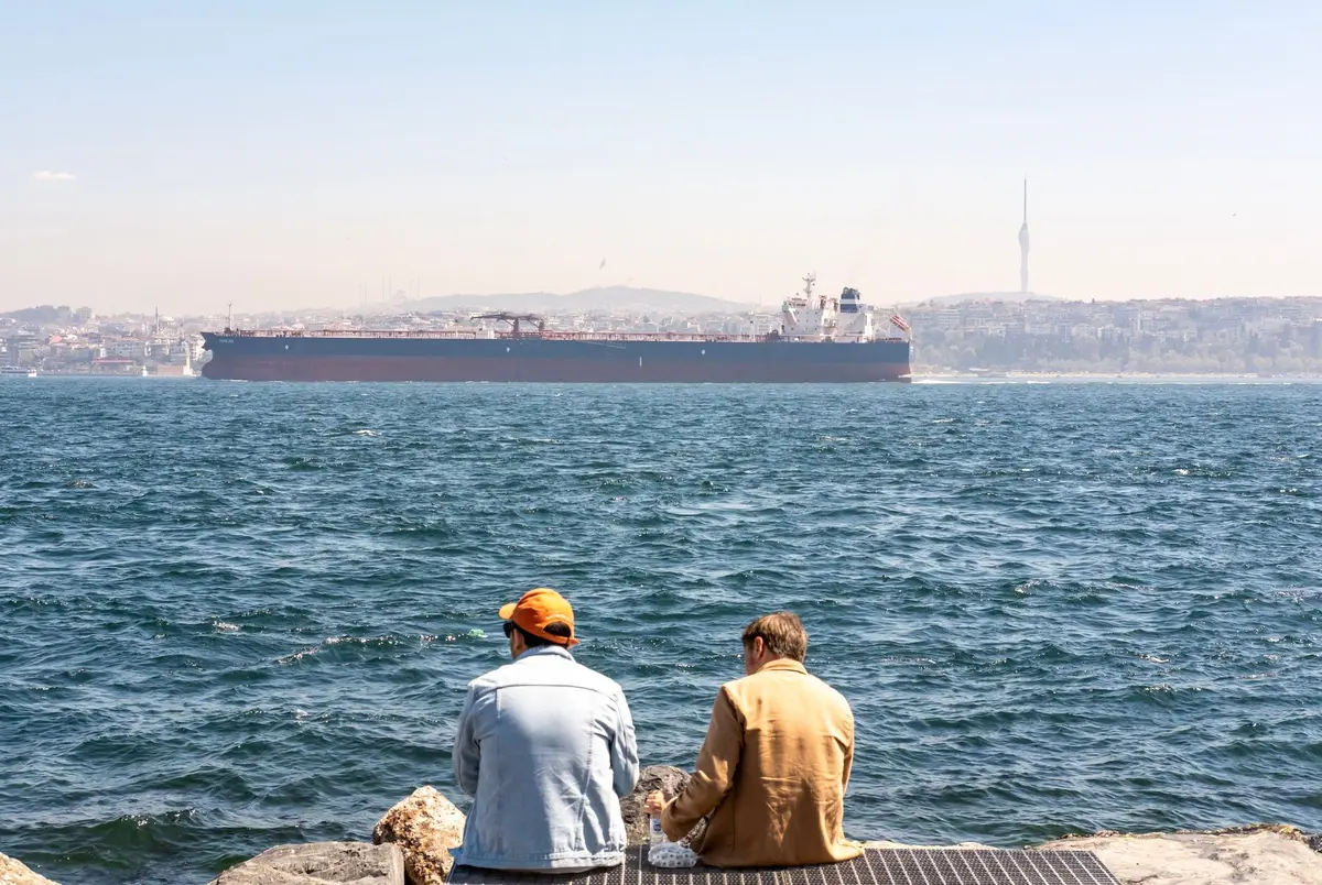 A wide view of the Strait of Hormuz with multiple commercial vessels anchored at a distance, minimal traffic on the waterway, a hazy horizon suggesting tension and restriction, one vessel with radar systems visible