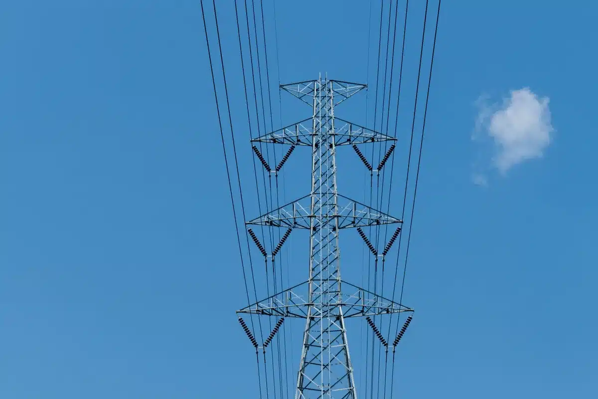 A high-voltage transmission tower silhouetted against a hazy sky with heat waves distorting the air, power lines stretching into the distance under extreme summer conditions