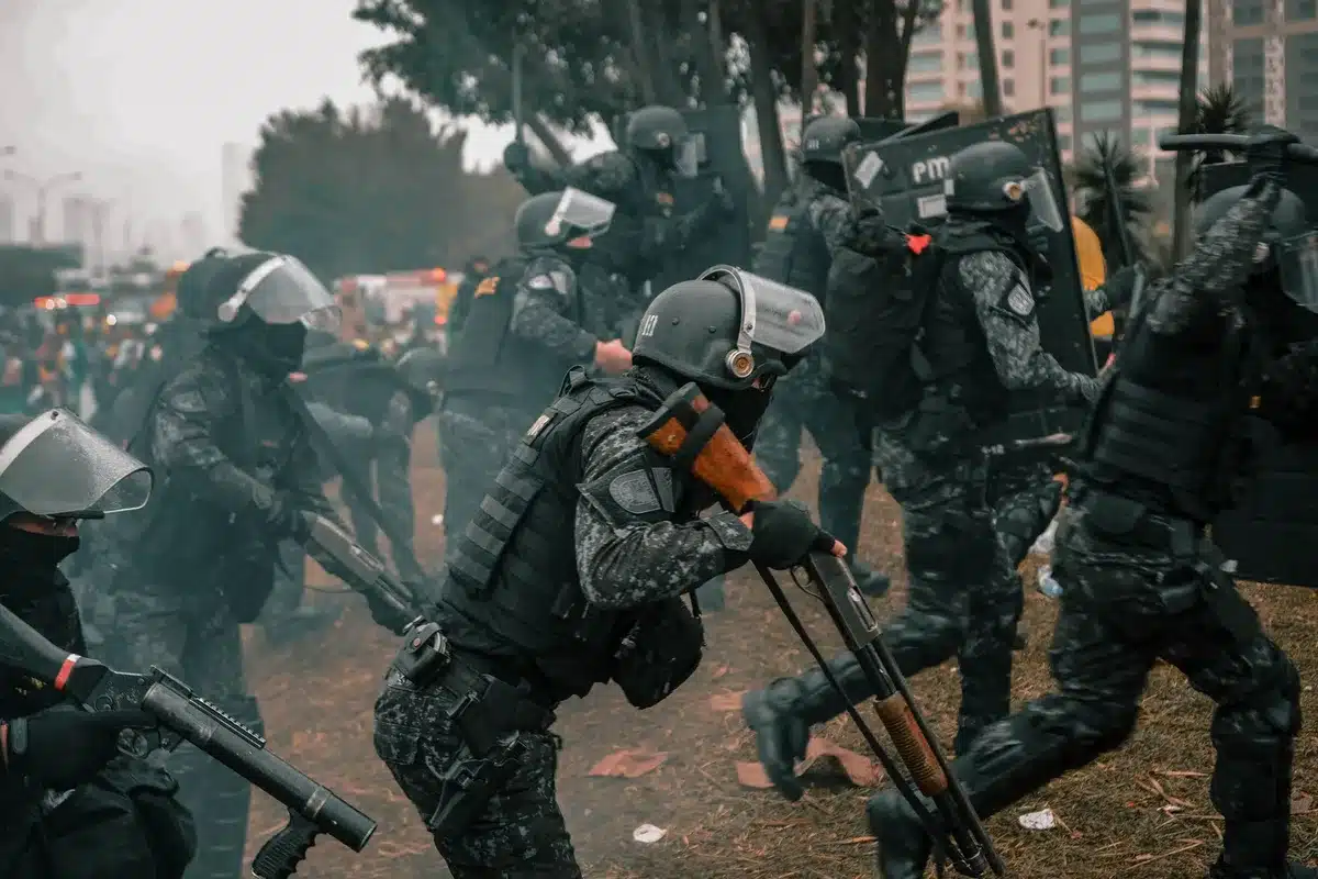 Police in riot gear standing with shields before a dispersing crowd, tear gas visible in the air, street pavement and urban buildings, a tense standoff captured mid-action