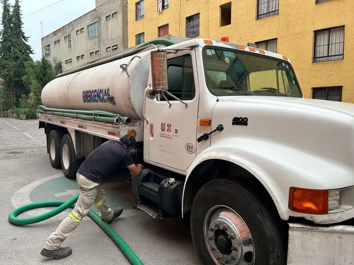 A water distribution truck dispensing from tanker into containers held by anonymous residents in a crowded urban plaza, afternoon light, faces obscured, collective strain visible