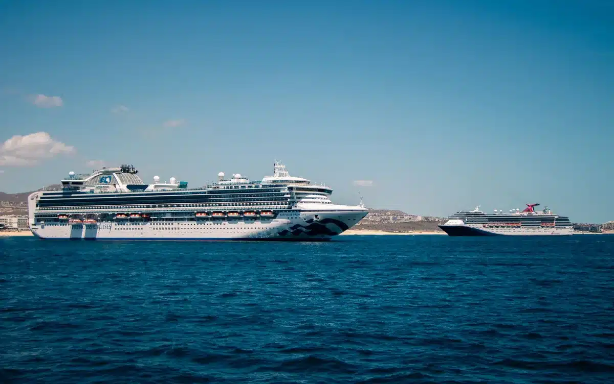 A modern cruise ship under bright sunlight sailing through calm ocean waters, multiple decks visible, passengers on upper deck areas, clear blue sky and water, sense of vacation leisure and travel