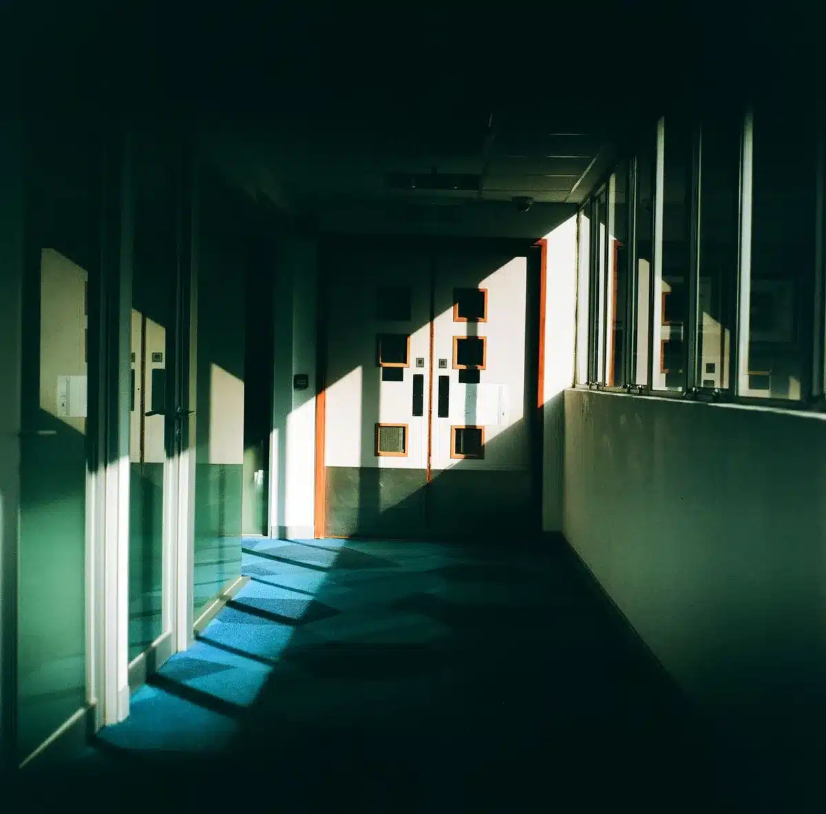 A dimly lit office corridor with one window showing daylight, empty chairs visible, shadows and light creating contrast, sense of solitude