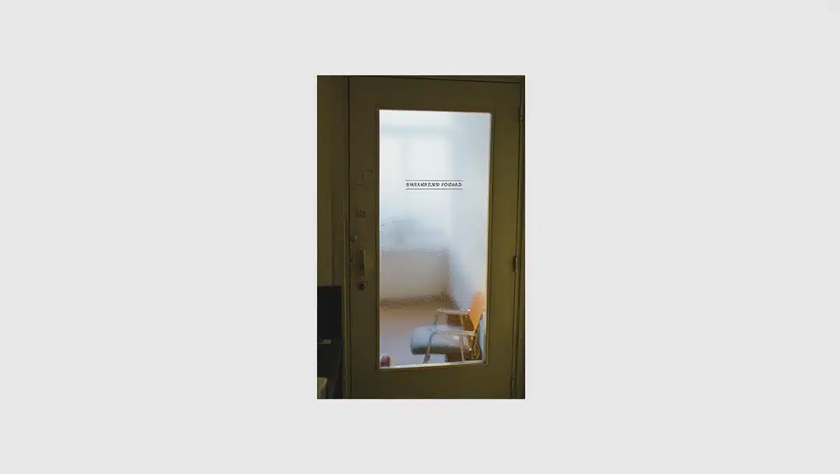 A closed office door with a nameplate removed, soft afternoon light casting long shadows across an empty desk visible through frosted glass | empty office aftermath
