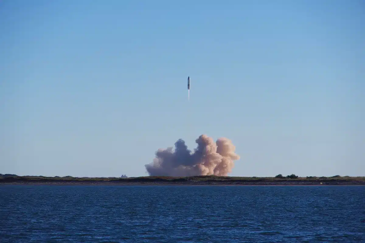 A Falcon 9 rocket ascending into the night sky from a coastal launch pad, bright orange flame illuminating the dark pre-dawn landscape, with the trajectory climbing at a steep angle against star-filled darkness.