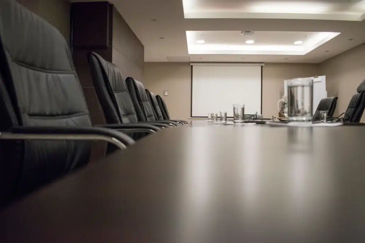 A corporate boardroom with an empty chair at the head of a long table, dim lighting casting shadows, documents and financial reports scattered, a window showing a grey sky, uncertainty and caution.