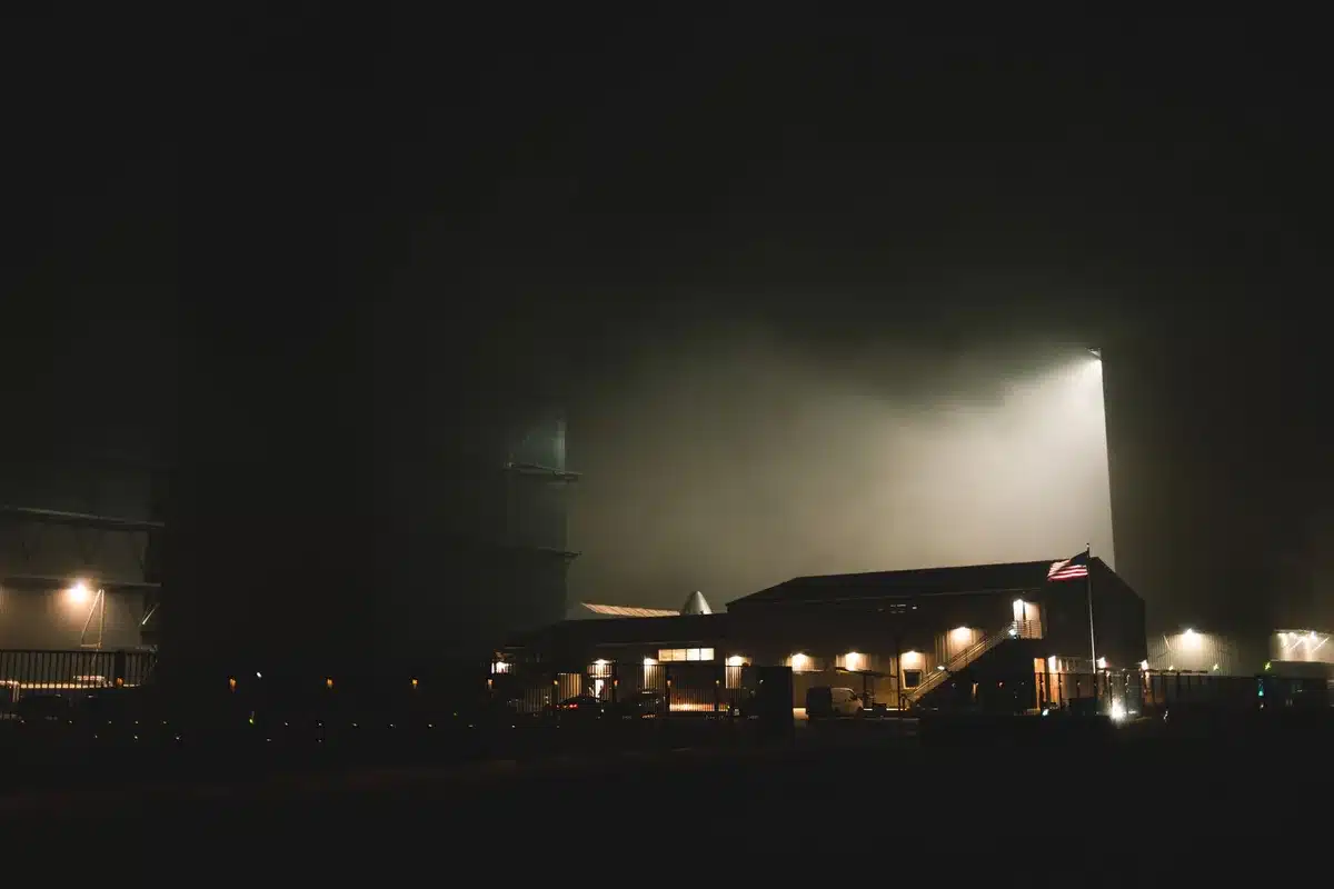 A tall stainless steel rocket on a launch pad at dusk, engines glowing with pre-ignition test fire, support structures and umbilical arms visible, storm clouds gathering on the horizon, tension and anticipation in the scene