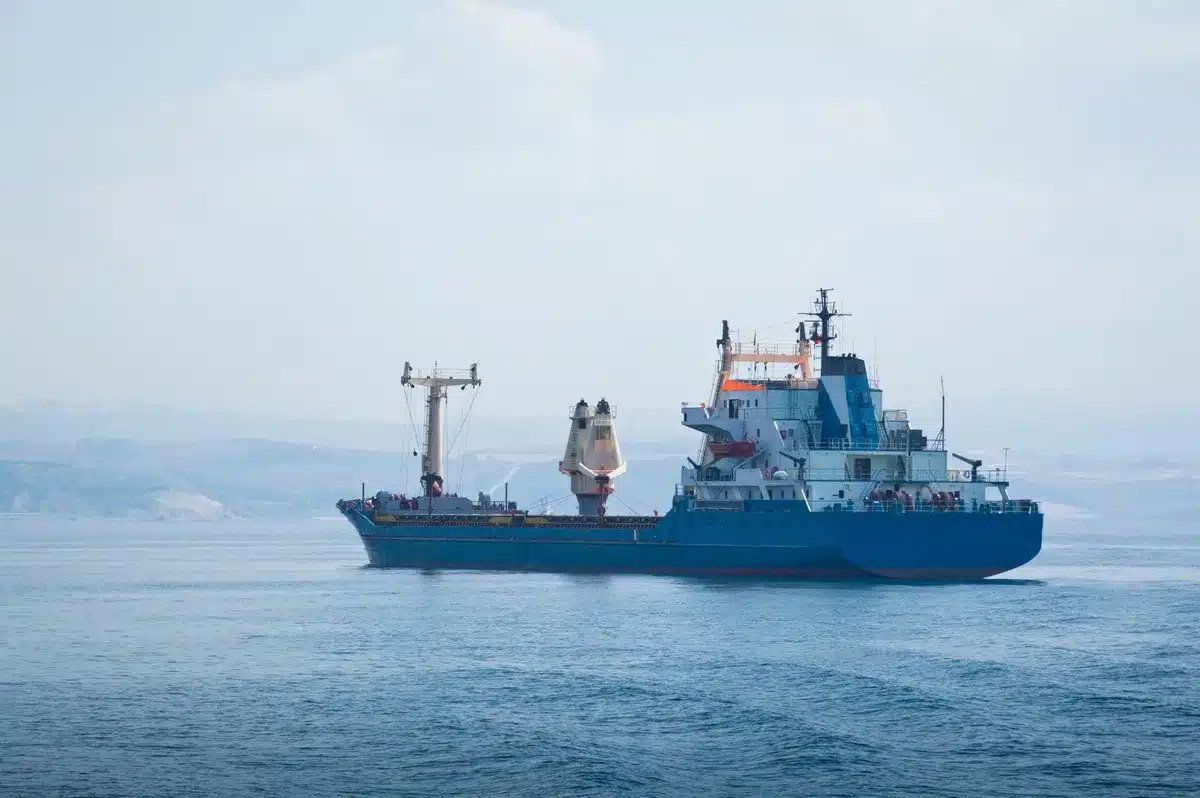A tanker ship with visible damage from a strike, listing slightly in calm waters, safety boat nearby, industrial port in soft focus behind, evidence of maritime conflict.