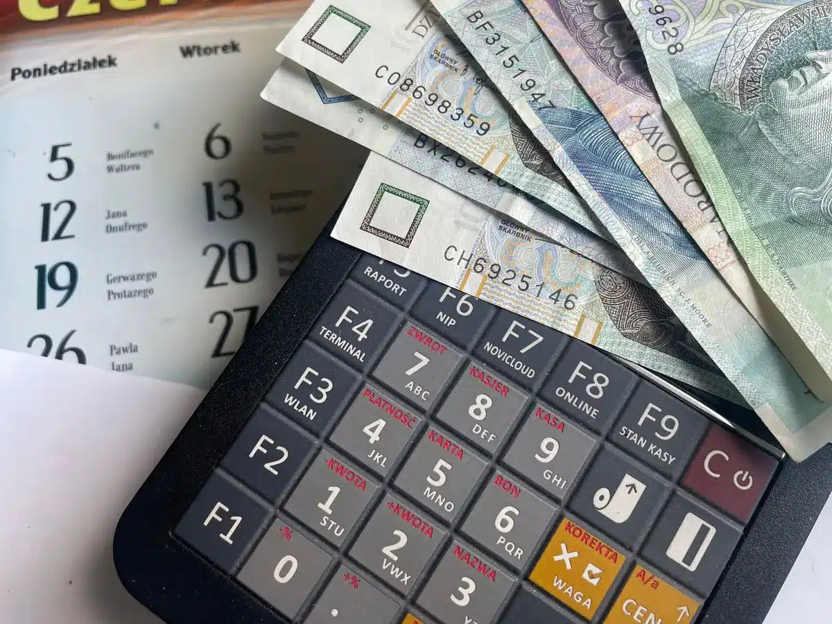 An older adult or person with a disability reviewing bills and financial documents at a table, with a calendar visible showing July and August dates, suggesting careful budget planning
