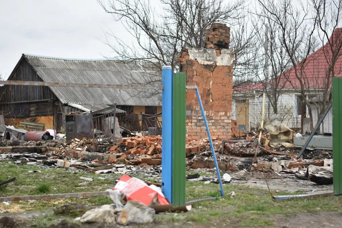 A destroyed industrial facility with collapsed walls and twisted metal beams, smoke still rising from rubble, daylight filtering through gaps in the structure, machinery buried under concrete and debris, one intact window frame standing amid the destruction.