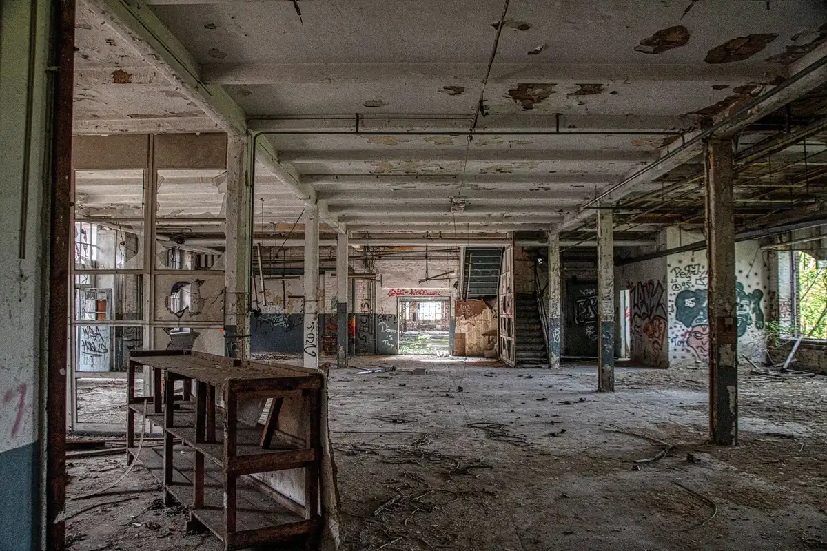 An empty factory floor with silent production equipment, workbenches covered in dust, overhead lights casting long shadows across the space, a single hard-hat resting on a bench, no workers visible, industrial silence.