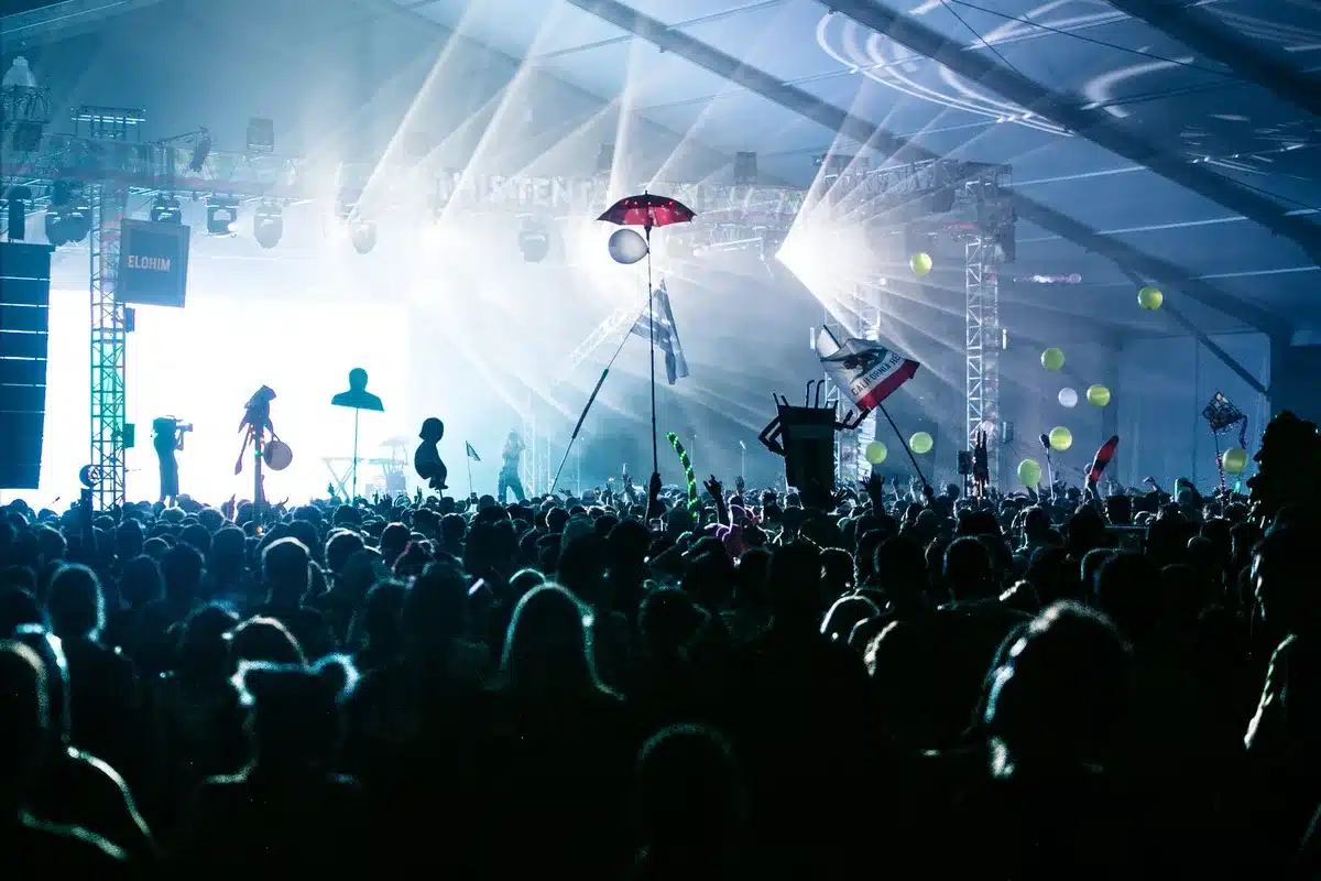 Concert venue filled with thousands of fans under bright stage lights, arms raised in unison, a sea of silhouettes against vibrant stage production, electric energy captured mid-performance